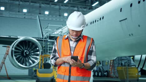 ingeniero masculino asiático con casco de seguridad usando un teléfono inteligente mientras está de pie con el avión en el hangar
