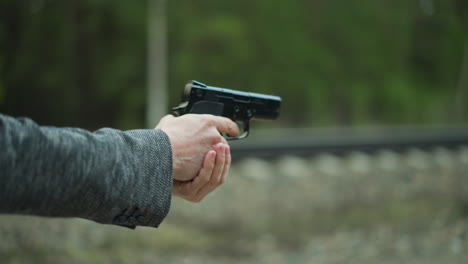 a close view of a person in a gray jacket drawing a handgun from a leather holster on their hip, the background shows a blurred railway track and forest