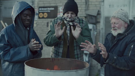 homeless men warming hands around fire barrel on cold urban street