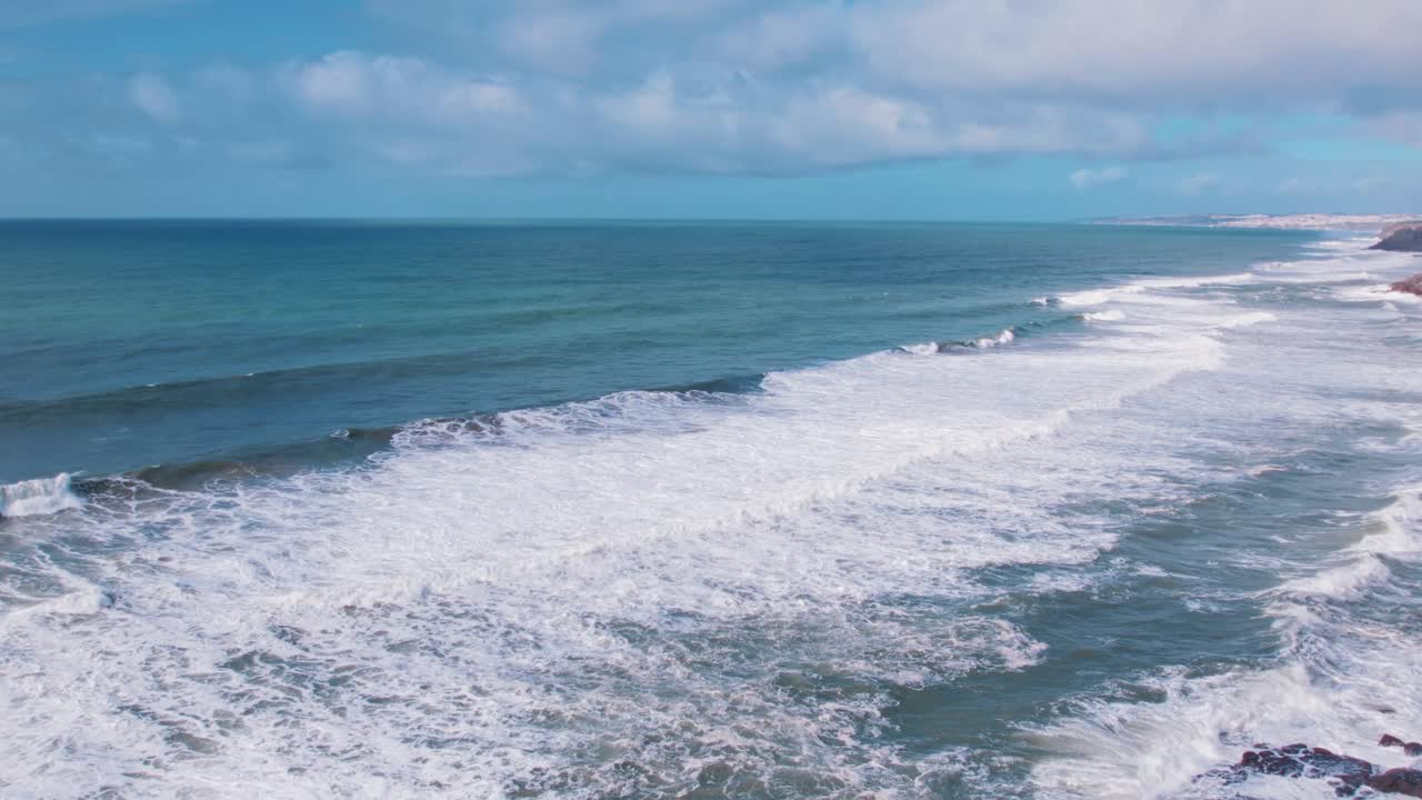 Aerial view of waves crashing against rugged cliffs near Ericeira, Portugal. Coastal greenery and foamy surf meet under a bright sky—capturing the raw beauty of the Atlantic shoreline