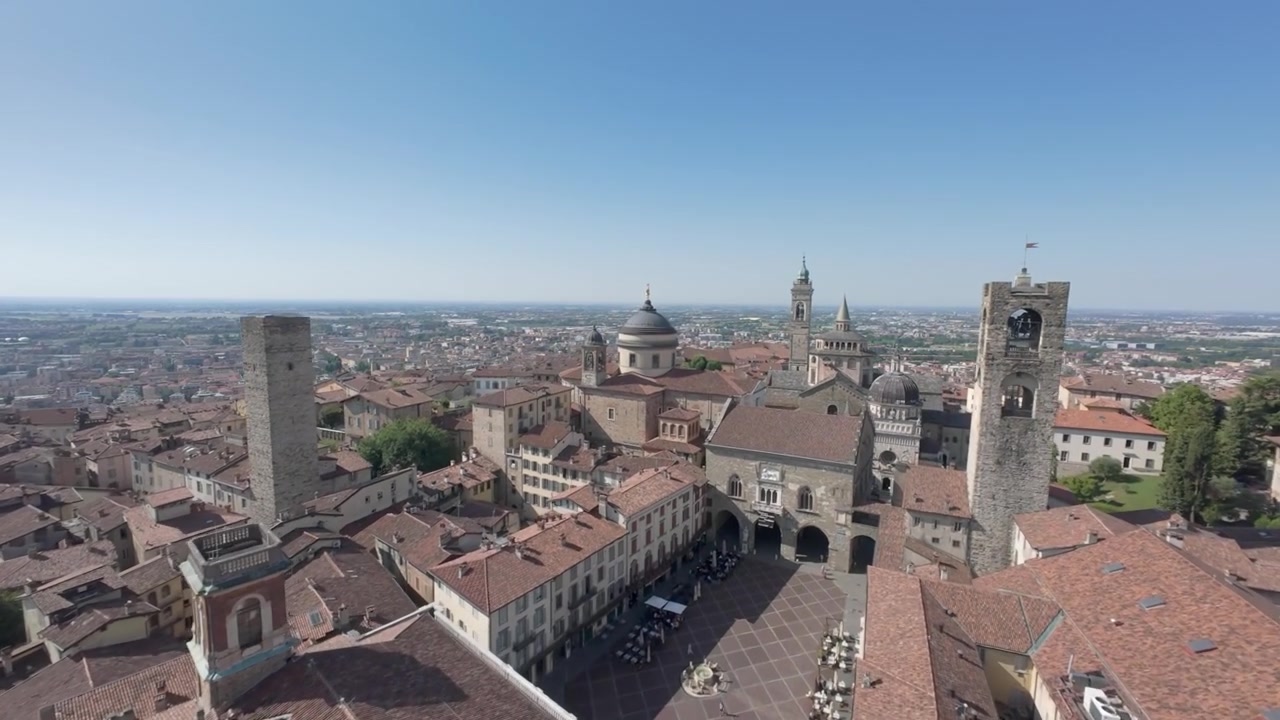 Un volo panoramico con drone che cattura una vista mozzafiato sui tetti storici, le torri e le piazze di Bergamo Città Alta sotto un cielo azzurro limpido.