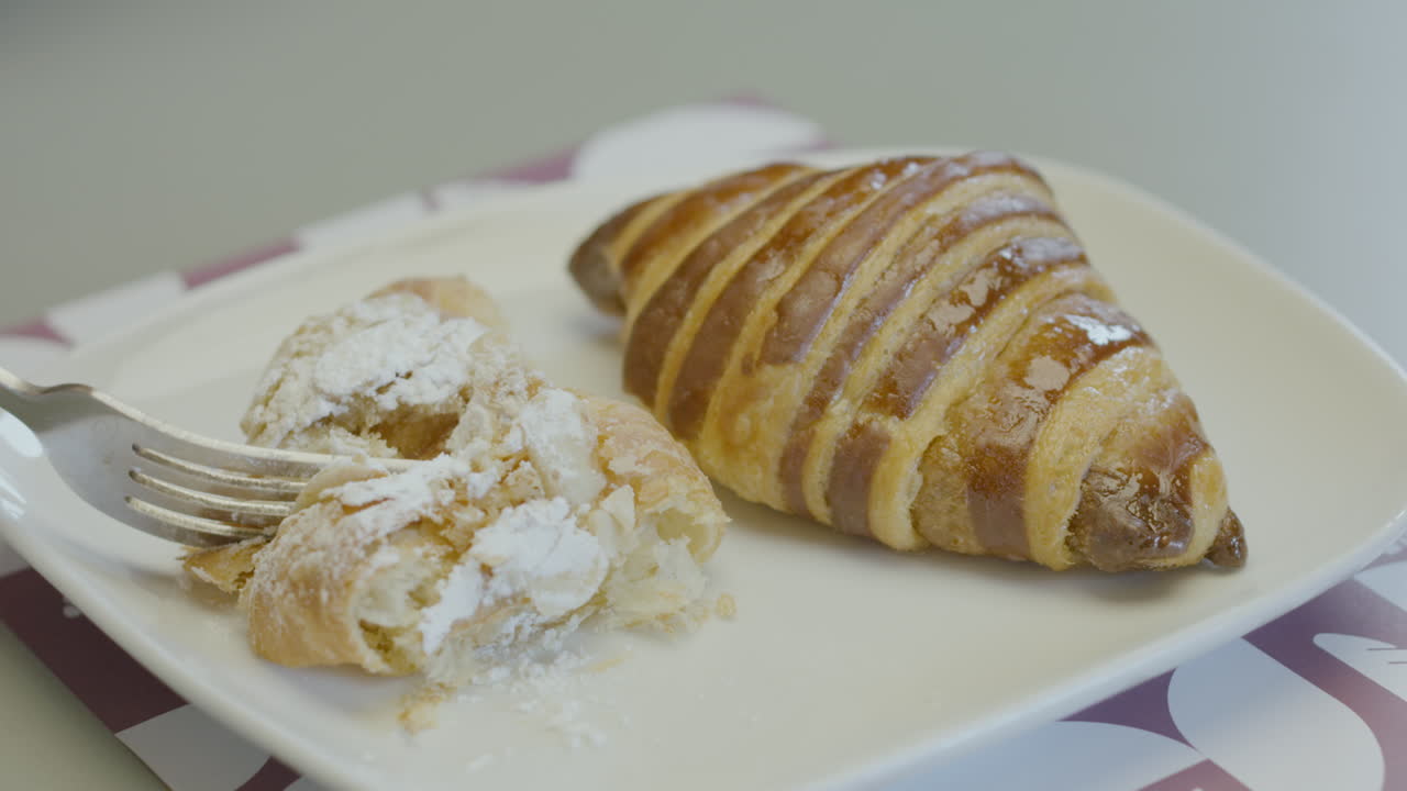 Primo piano di un dolce croissant glassato e cosparso di zucchero a velo, mentre una forchetta ne taglia una porzione mostrando la sua consistenza croccante e morbida