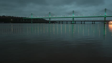 A-lit-bridge-reflects-on-a-calm-river-at-twilight-under-a-cloudy-sky