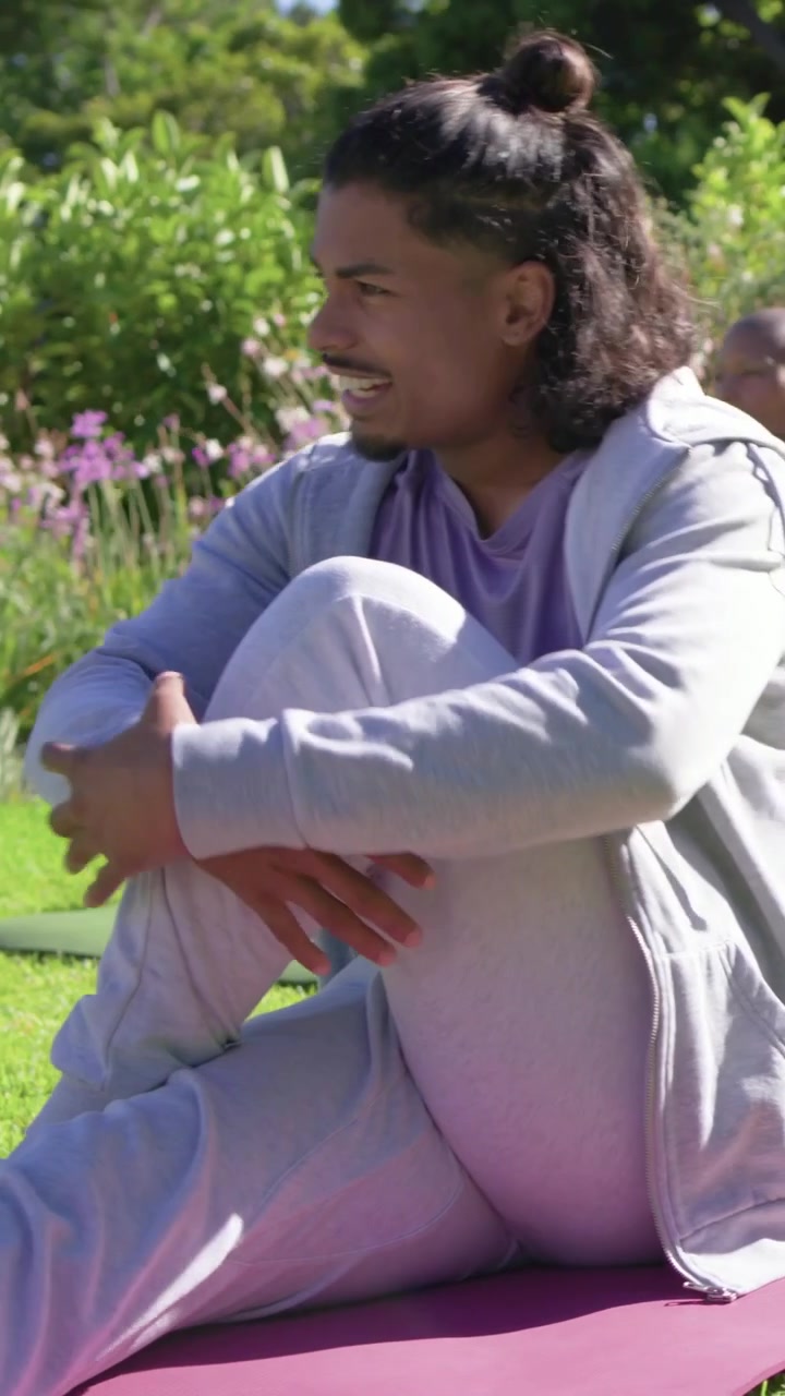 Vertical video: Camera shifting, man in gray top pulling knee turning speaking for yoga on pink mat