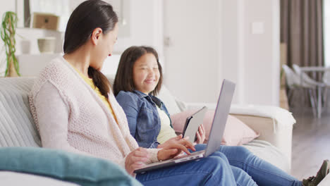 video of happy asian mother and daughter sitting on sofa with laptop and tablet