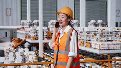vista lateral de una ingeniera asiática con casco de seguridad hablando en un teléfono inteligente mientras está de pie en el almacén con estantes llenos de productos de entrega
