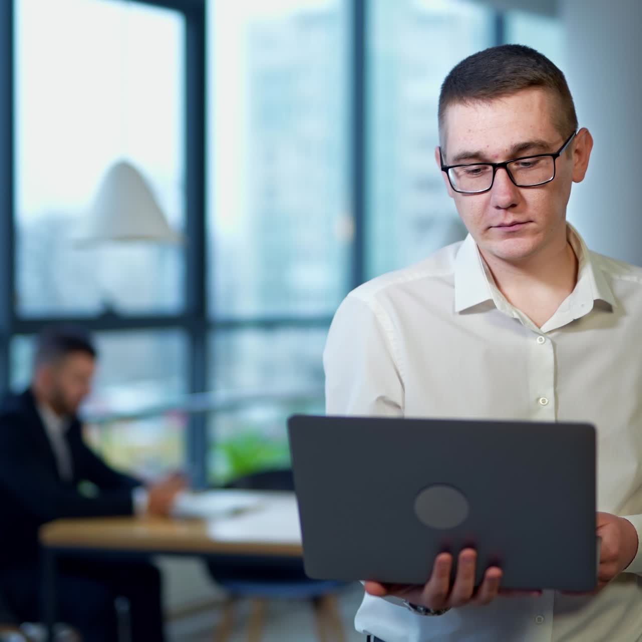 Male manager with laptop in one hand stands in office. Young man describes something on computer. Man in suit in blur at backdrop