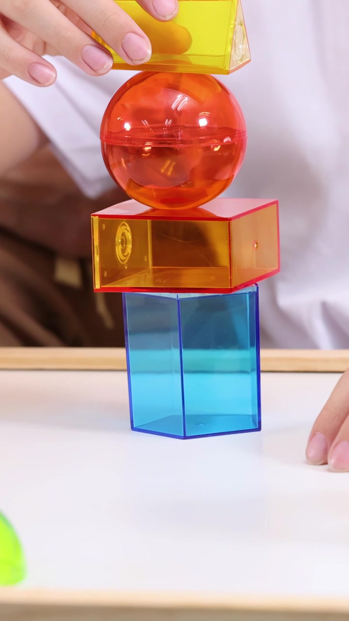 Close-up tangan anak menyeimbangkan balok geometris transparan di atas meja dalam pencahayaan kelas yang lembut.