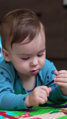 caucasian toddler lies on the floor looking carefully at the black lace. kid turns his head to camera watching it intently. toys around baby. vertical video