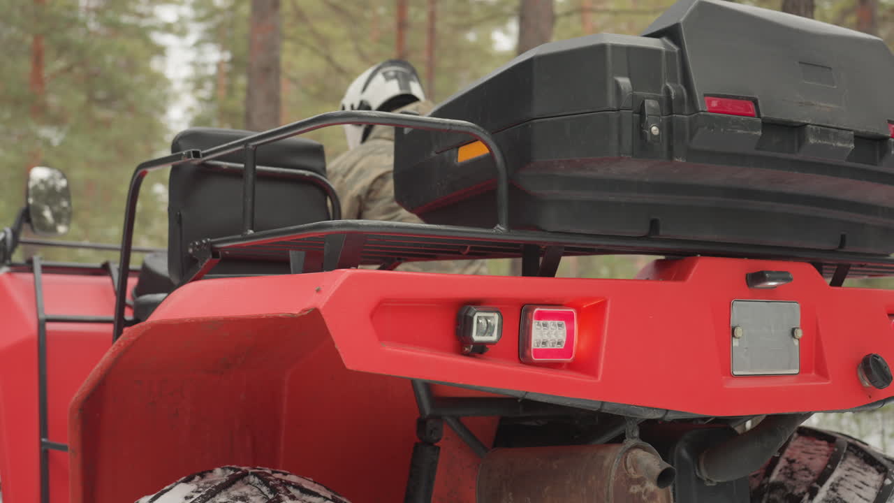 I primi piani del posteriore di un ATV mostrano il fanalino e la marmitta sotto il quad rosso. Il pilota esegue una rapida manutenzione in una pineta innevata: scarico congelato, pneumatici macchiati di neve, robusto baule portaoggetti e portapacchi posteriore, mentre prepara il veicolo.