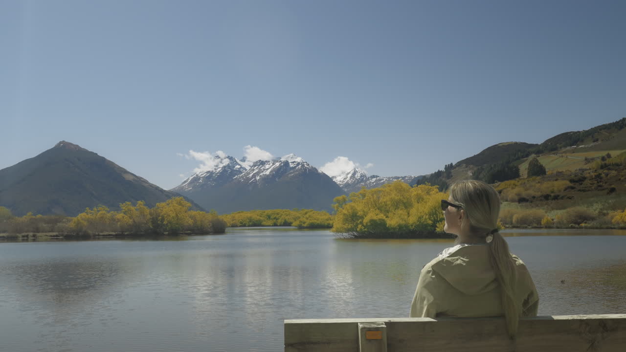 글레노키 석호의 햇살 가득한 전망을 즐기며 나무 벤치에 앉아 있는 금발 여성 관광객