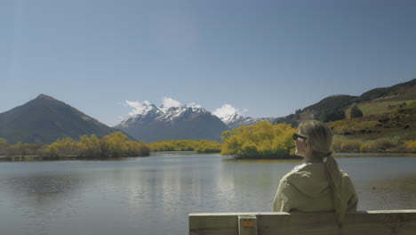 Blonde-Touristin,-Die-Auf-Einer-Holzbank-Sitzt-Und-Einen-Sonnigen-Blick-Auf-Die-Lagune-Von-Glenorchy-Genießt