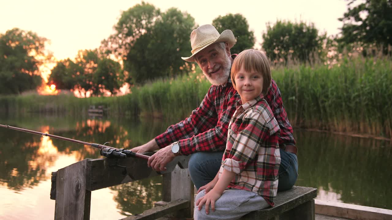 Close-up view of a grandfather and his grandson fishing and talking sitting on the lake pier on a summer day at sunset