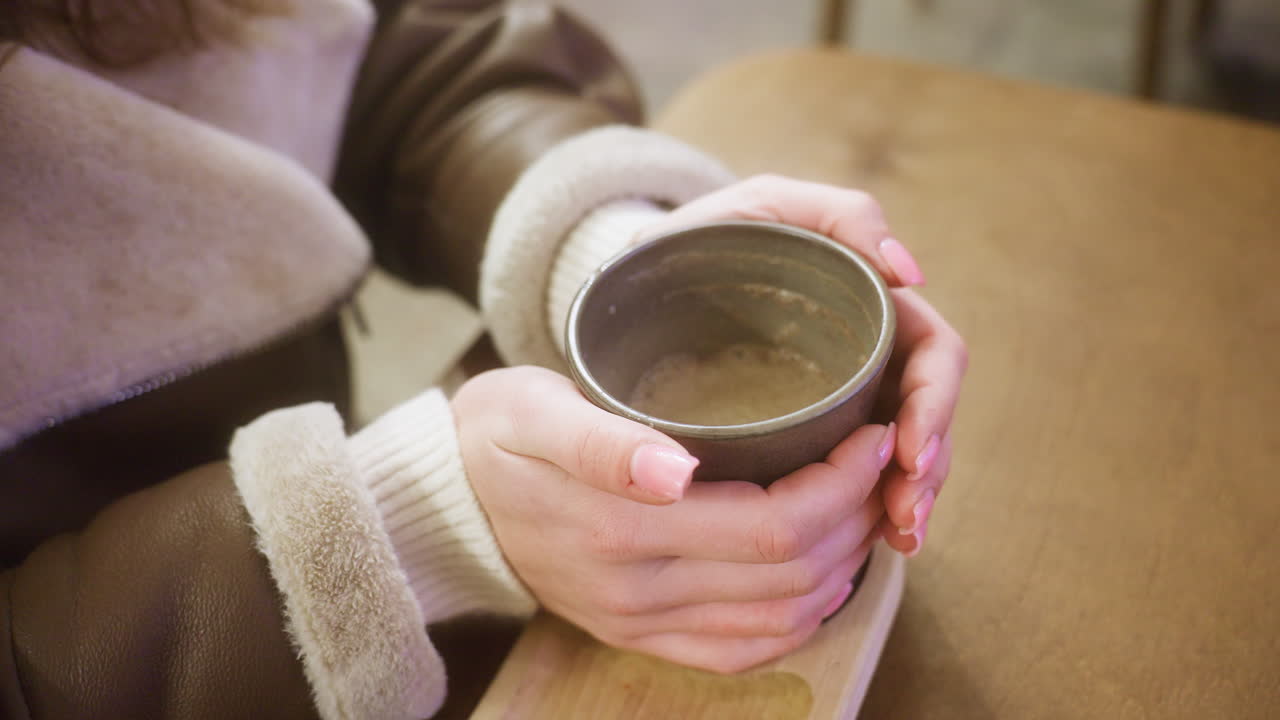 Close-up shot of woman's hand in brown shearling jacket, holding a cup of coffee, gently lifting it at cozy cafe. Warm ambiance and soft lighting create relaxing atmosphere perfect for enjoying hot drink