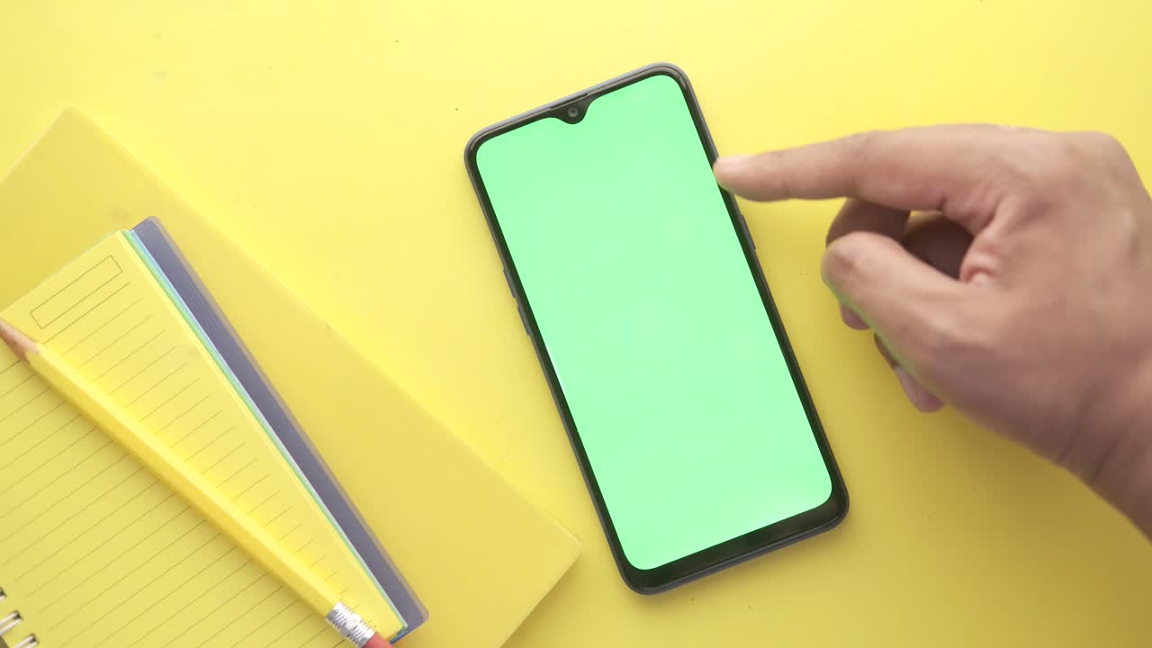 mano tocando un teléfono inteligente de pantalla verde en un fondo amarillo con cuaderno y lápiz