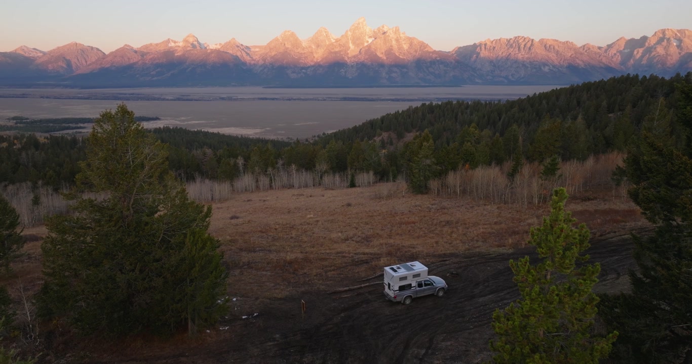 RV camper parked on dirt overlook with Grand Teton mountains at sunrise, aerial tilt-up