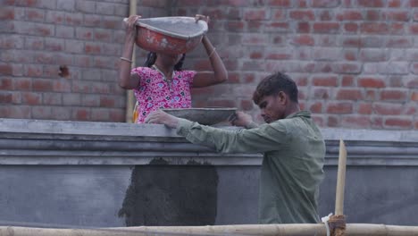 a slow motion handheld long wide shot of an indian woman wearing pink kurta and a man wearing faded olive green shirt, working at a construction site, woman passing liquid concrete from a tub