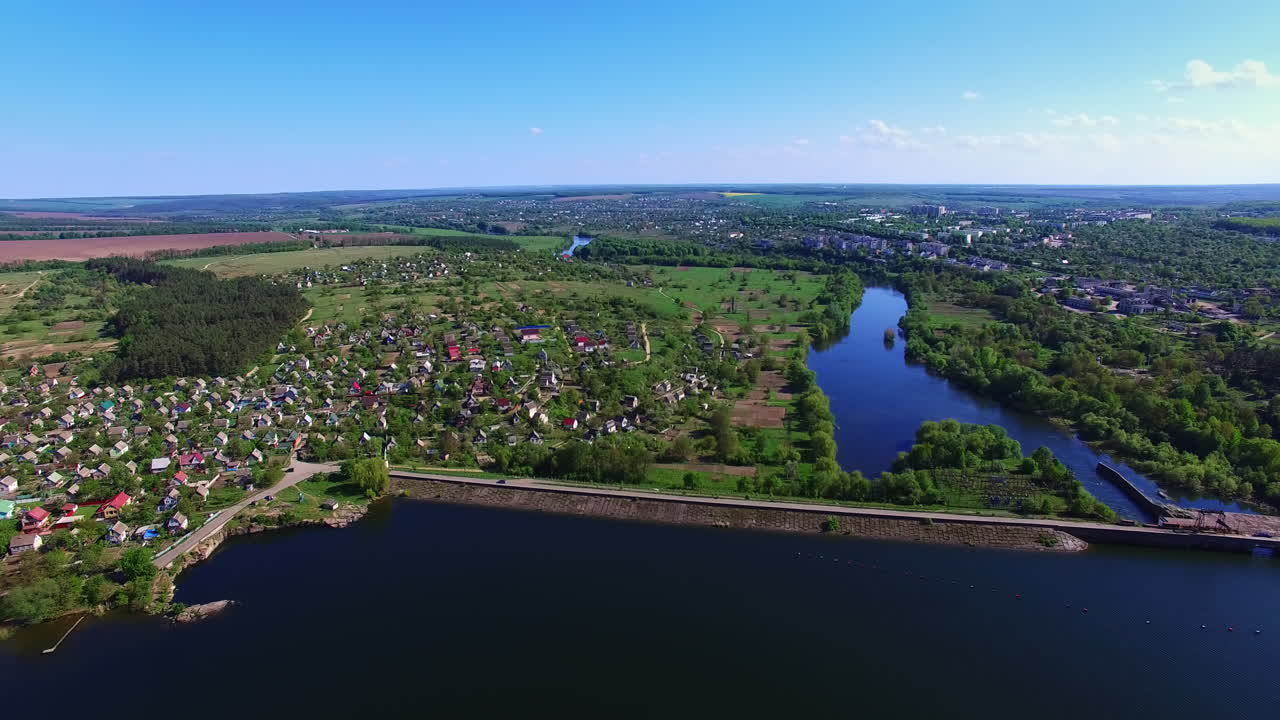 Approaching the beautiful village dwelling on the bank of blue river. Green landscape under clear sky at backdrop. Top view.