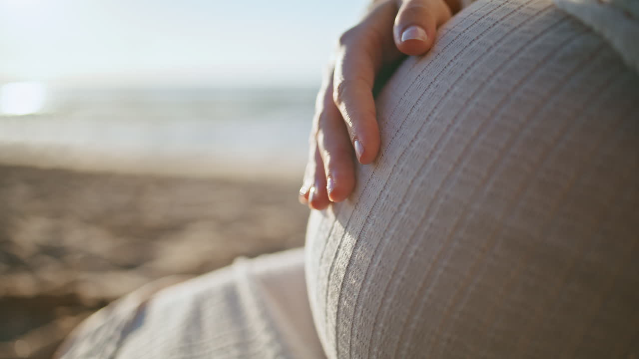 미래의 엄마가 해가 지는 해변에서 배를 만지며 여성 손이 배를 만지고 있습니다.