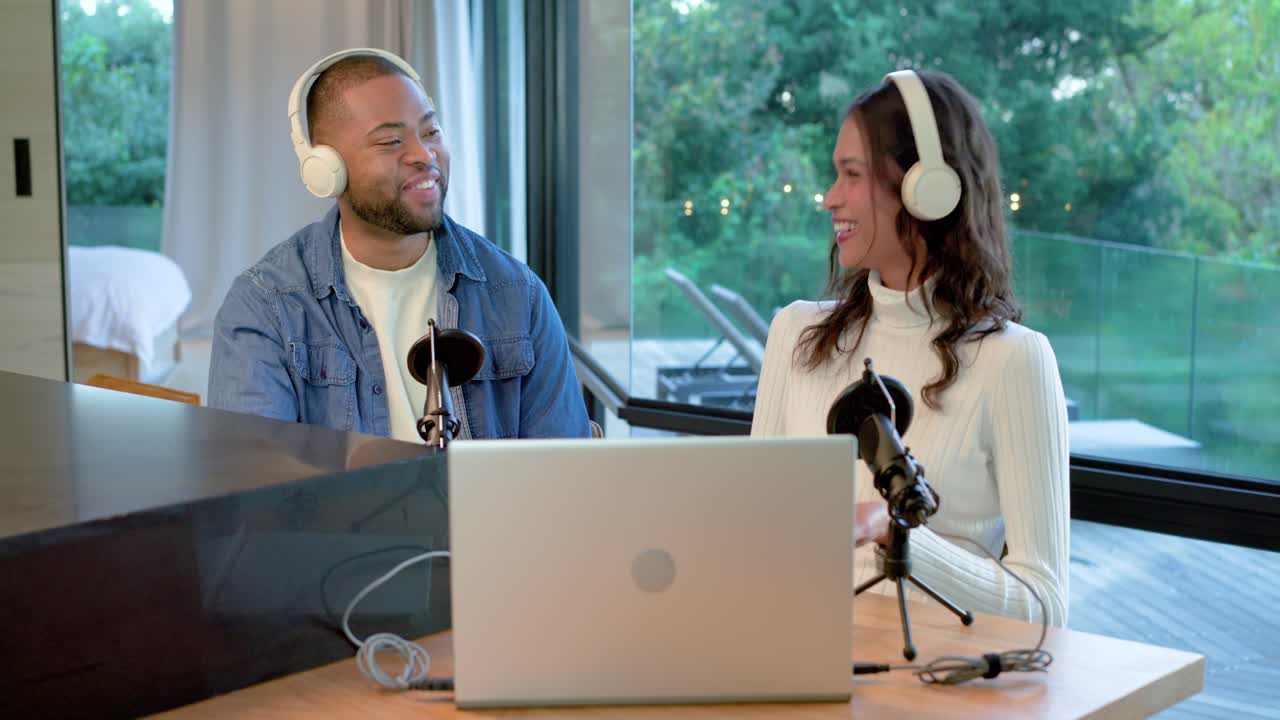Diverse co-hosts leaning, gesturing and typing on laptop into microphones while wearing headphones