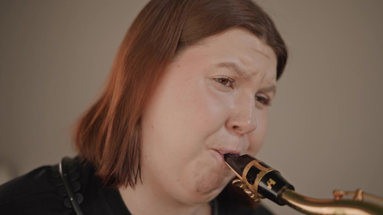 Femme jouant du saxophone en gros plan en studio, cours privé axé sur la sonorité et l'embouchure, détail du bec doré, fond brun doux, exercices d'improvisation et d'échauffement, concentration intense