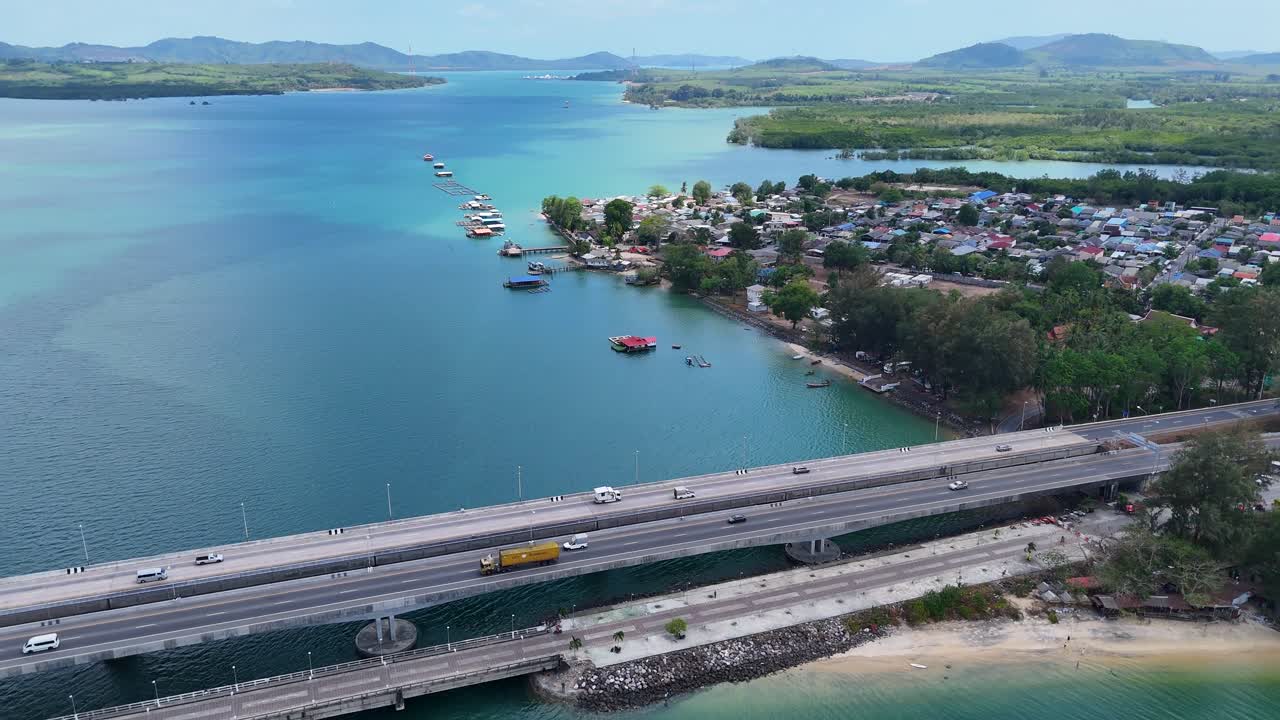 드론 영상은 태국 푸켓의 사라신 다리를 포착합니다. 푸른 하늘 아래 푸른 푸른 물과 멀리 있는 산을 가로질러 펼쳐져 있습니다.