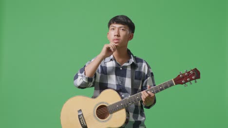 asian teenager with guitar thinking about something and looking around then raising his index finger on green screen background