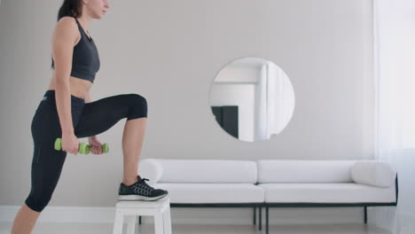 close-up young beautiful brunette woman steps on a chair with dumbbells in her hands.