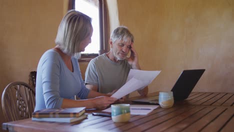 senior caucasian couple using laptop and holding documents at table