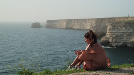 mujer sentada en un acantilado con vistas al mar usando su teléfono