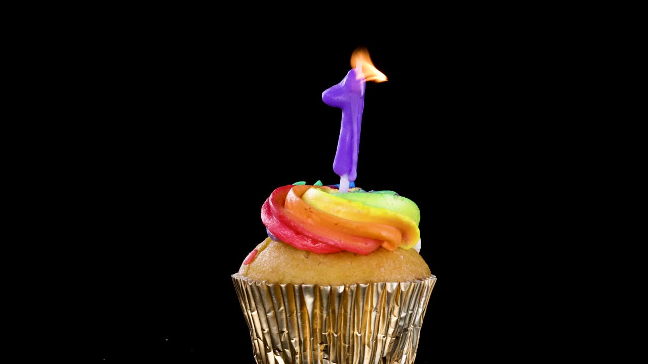 A single cupcake with rainbow frosting and a lit purple number one candle, isolated on a black background, brightly illuminated with no visible movement