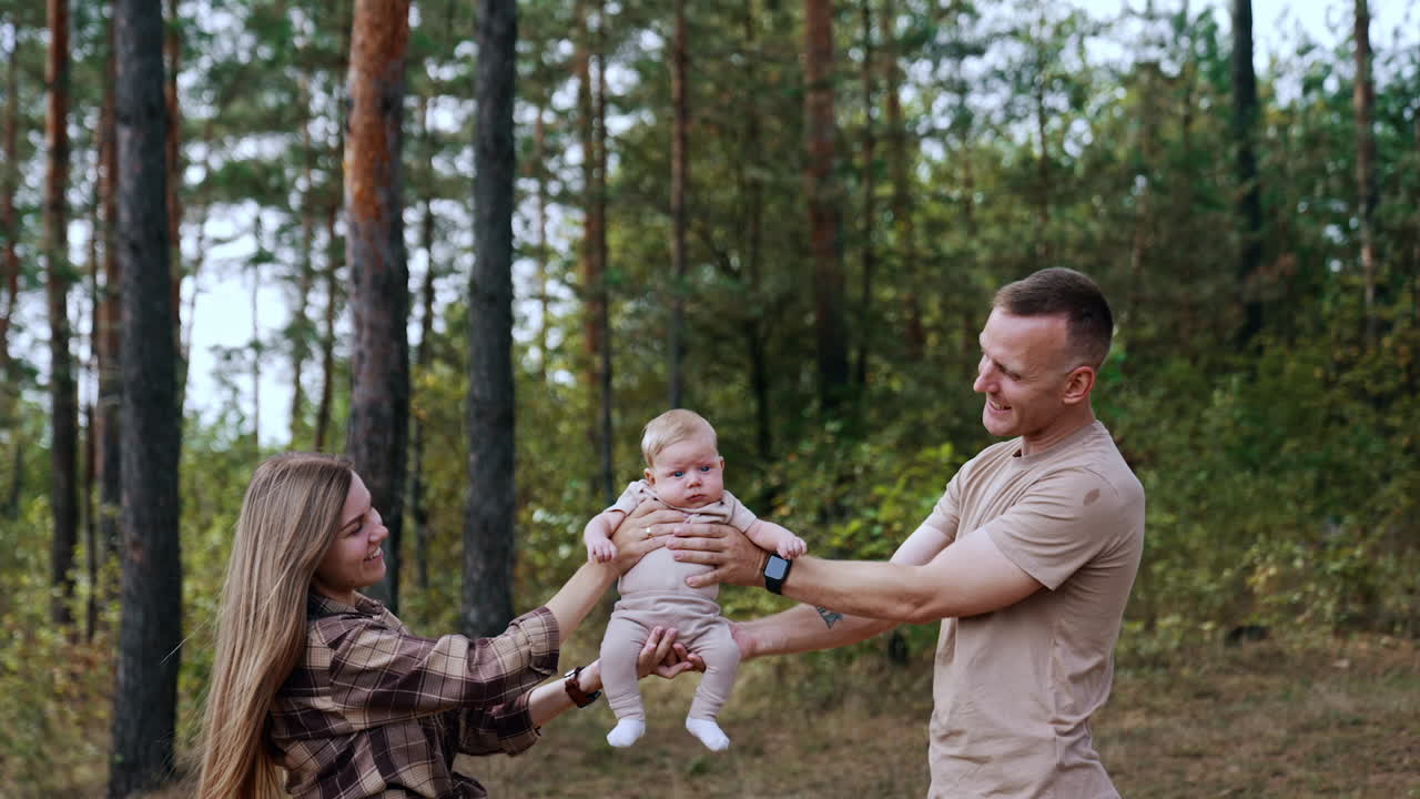 First time parents holding their son from two sides. Little kid sits peaceful and calm in mom's and dad's hands. Nature at backdrop.