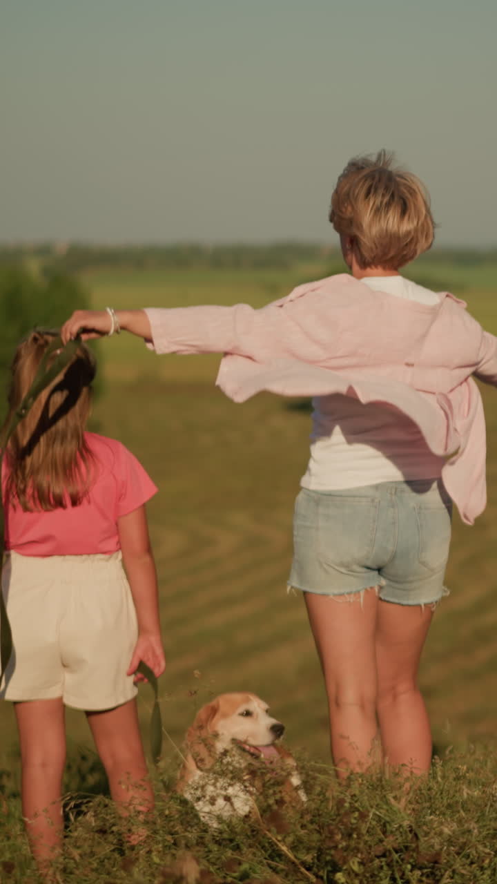 은 하늘 아래 언덕의 농지 위에 서 있는 가족의 뒷면 모습, 어머니가 딸에게 멀리서 흥미로운 것을 가리키고, 다른 여성이 꽃을 들고 배경에 서 있습니다.