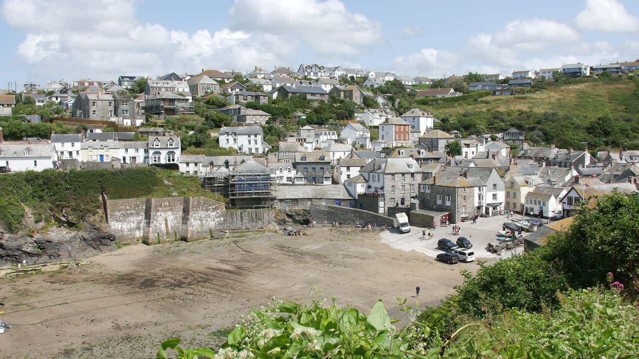 En havn i Port Isaac, England, med fiskebåde på grund i den sandede bugt ved lavvande, indrammet af robuste klipper og det klare, blå Atlanterhav. Portwenn kystmure og tidevandsbassiner