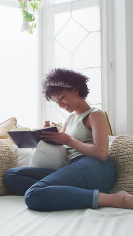 vertical video of happy biracial woman relaxing at home, writing in notebook, with copy space