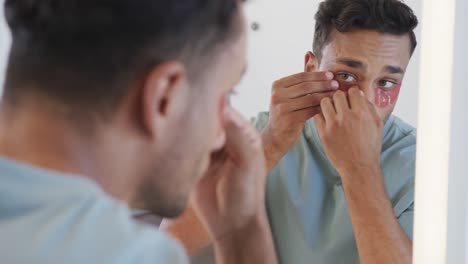 Biracial-man-looking-in-mirror-applying-under-eye-masks-in-bathroom,-slow-motion