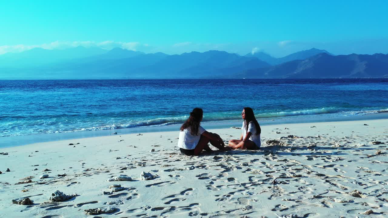 Two girls sitting on white sand of exotic beach talking about their vacations, in front of blue sea bordered by high mountains of Indonesian islands