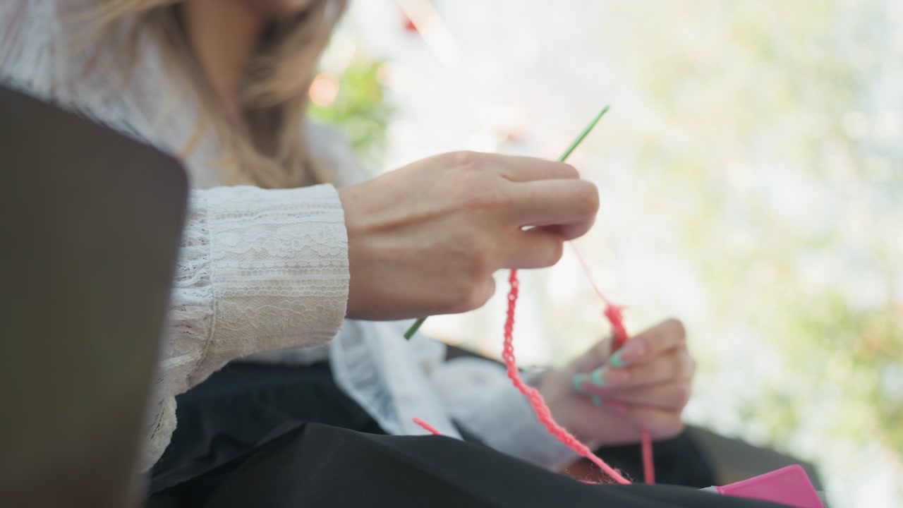 Primo piano di una giovane donna che lavora all'uncinetto con filato rosa brillante e uncinetti verdi, indossa un maglione di pizzo bianco. Un laptop è parzialmente visibile sul lato sinistro in un ambiente con luce diurna soffusa. Sfondo sfocato.