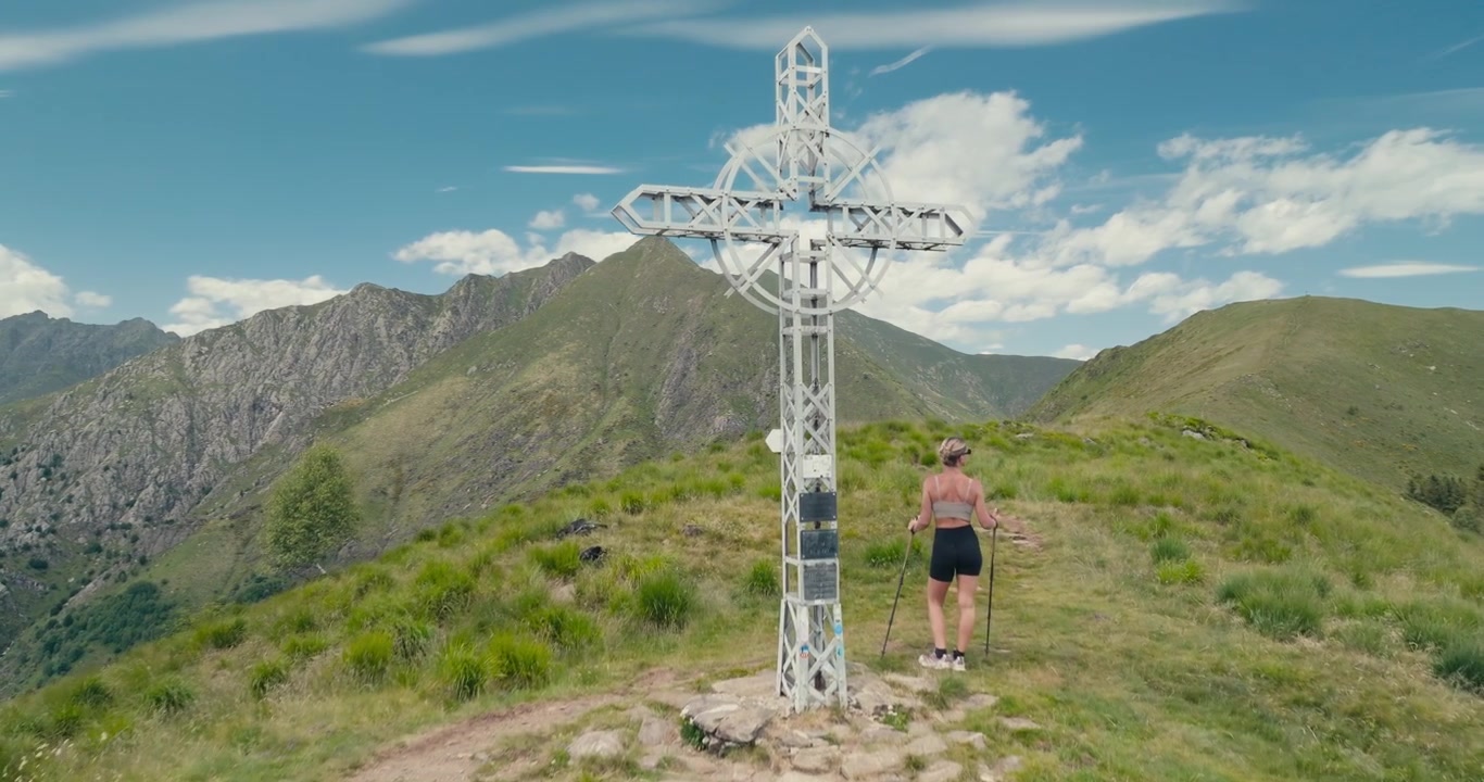 En drone flyver over træer mod et topkors, mens en kvindelig vandrer går de sidste meter til toppen, omgivet af betagende bjergudsigter under en klar blå sommerhimmel.