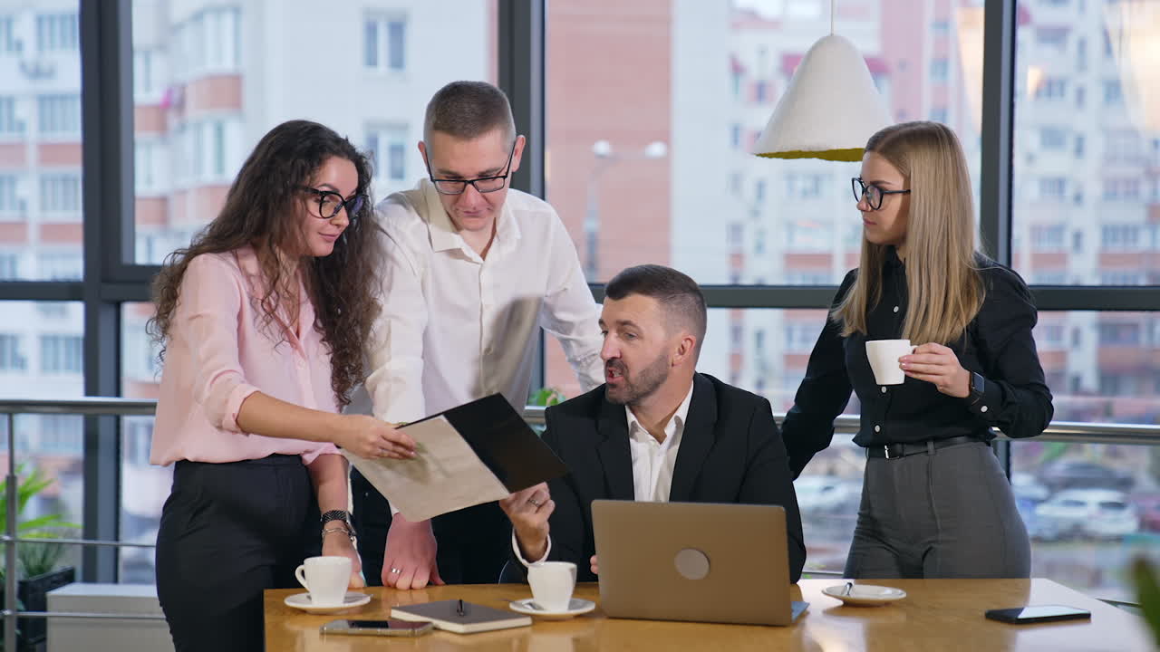 Team of colleagues look at documents and talk about them. Boss sitting at the table listens to his employees, responds to them and smiles.