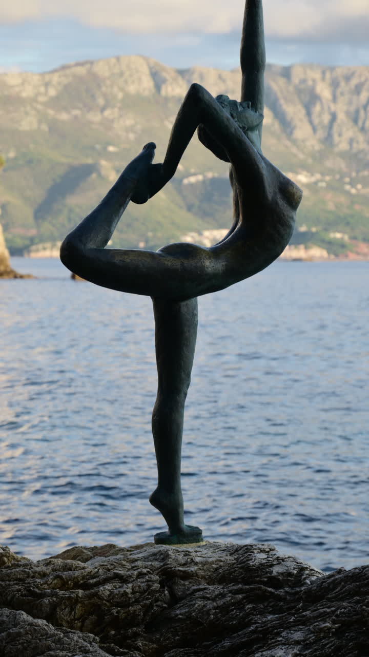 Estátua de dançarina de bronze de pé junto ao mar em Budva, Montenegro, com a arquitetura da cidade antiga ao fundo.