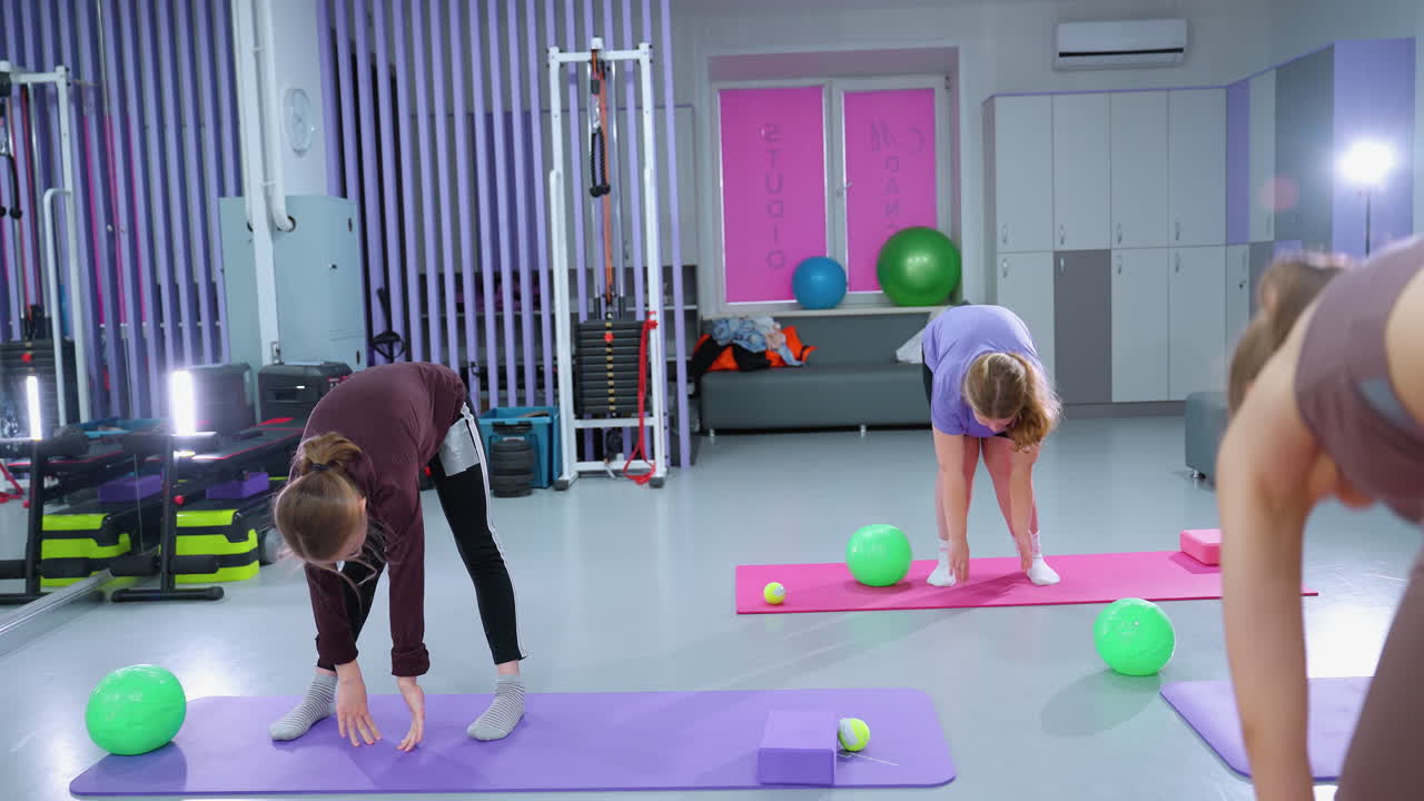 체육관에서 앞으로 구부러지는 운동을 통해 어린 소녀들을 안내하는 강사의 부분적인 시각, 참가자들은 유연성과 균형에 초점을 맞추며 팔을 펴고 움직임을 따르고 있습니다.