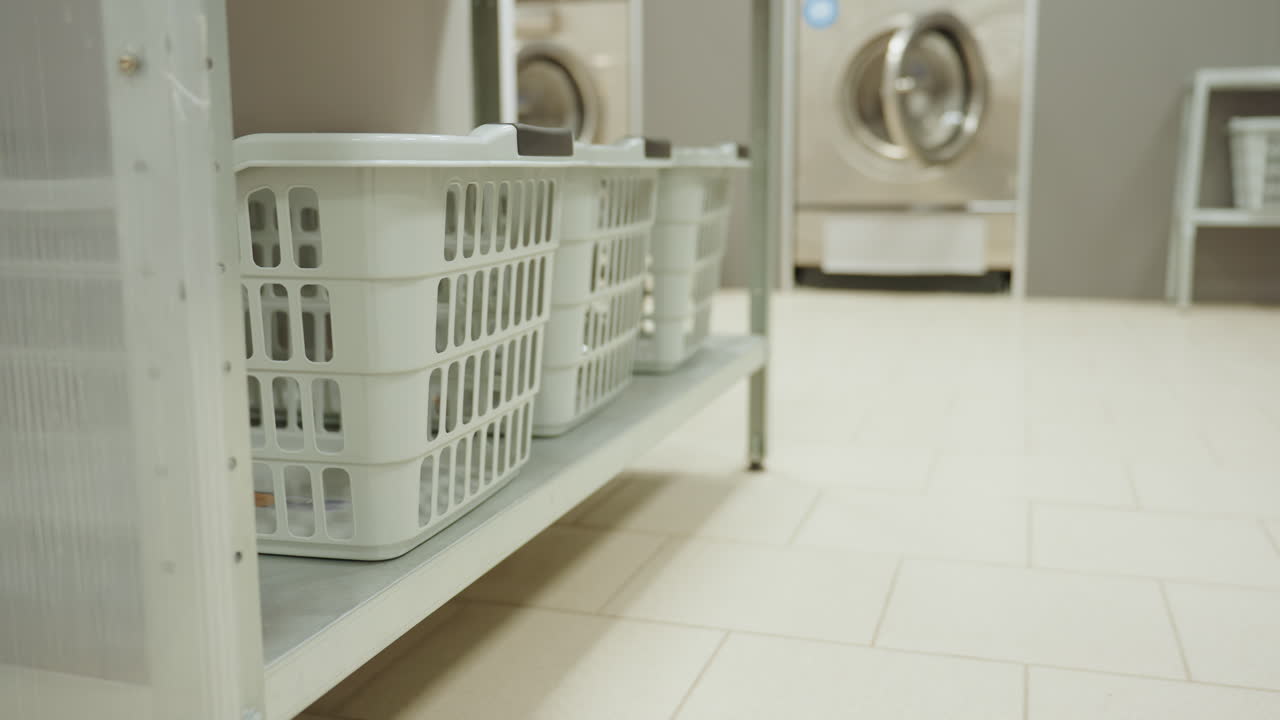 Side view laundry baskets on metal shelf in blurred laundromat interior, stainless machines and tiled floor in soft focus, organized clean utility storage detail for service and maintenance