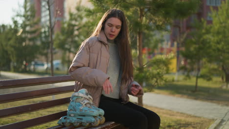 a young girl wearing a peach jacket, grey inner shirt, and black trousers sits on a park bench. she places her rollerblades beside her and rests her hands on her chin