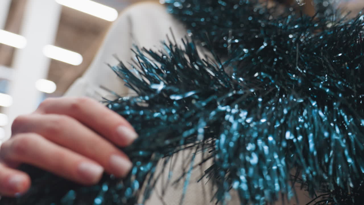 Close-up of person gently feeling texture of dark blue tinsel, with shimmering festive decor and blurred background creating a warm, magical holiday shopping experience in decoration store