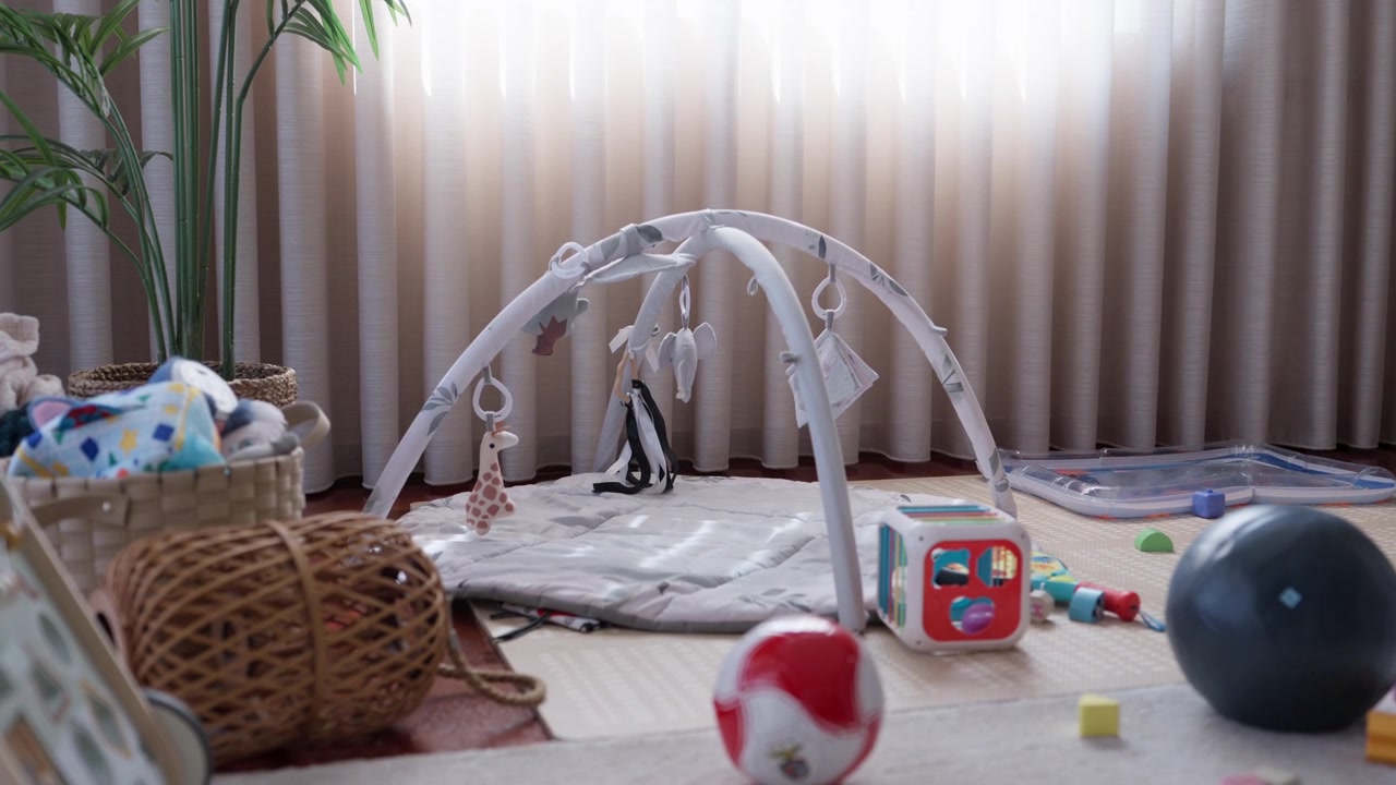 リビングルームの床に置かれた、吊り下げおもちゃのボールとアクティビティキューブが付いたベビープレイジム。カーテンのそば。