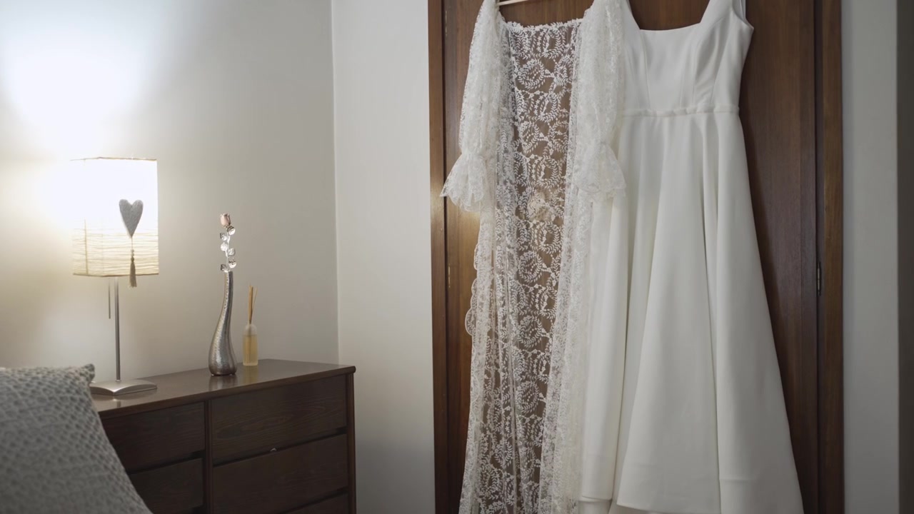 Abito da sposa bianco appeso alla porta dell'armadio accanto al comodino e alla lampada.