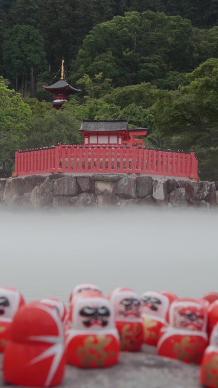 Video verticale che mostra bambole Daruma colorate in primo piano con una tradizionale struttura del tempio rosso che emerge attraverso la nebbia e la vegetazione lussureggiante al tempio Katsuoji a Osaka, in Giappone
