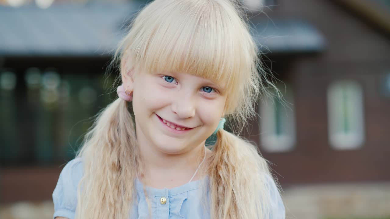 Porträtt av en blond flicka som ler mot kameran. Hon står i bakgrunden av sitt hus. Video med kort skärpedjup.