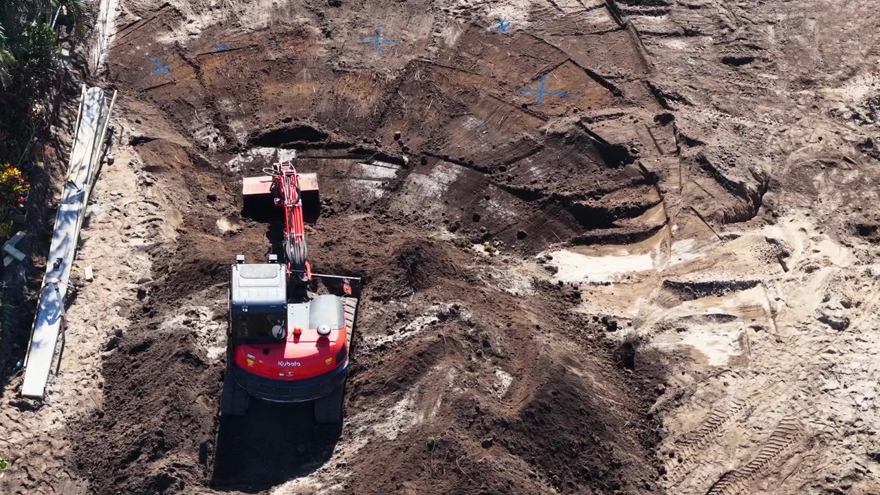 High angle shot of compact excavator digging and leveling dark earth at urban construction site.
